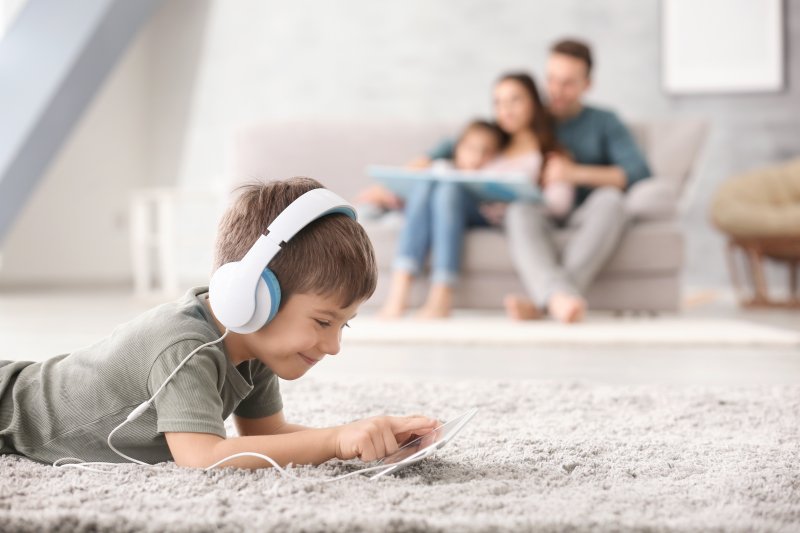 a little boy wearing headphone and playing on a tablet while lying on the carpet